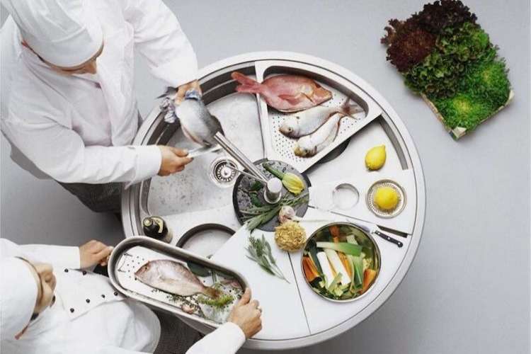 Two chefs wearing white chef gear working on a round chrome work space