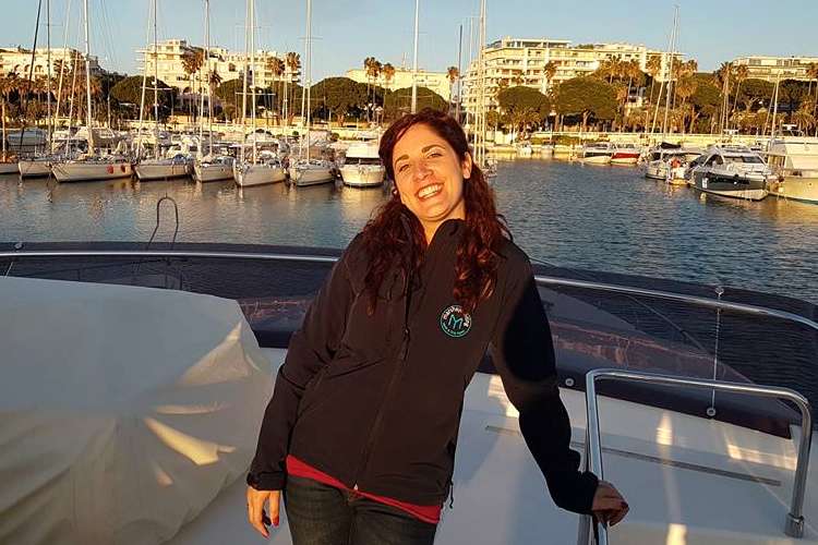 Woman standing on a yacht deck with a port in the background.